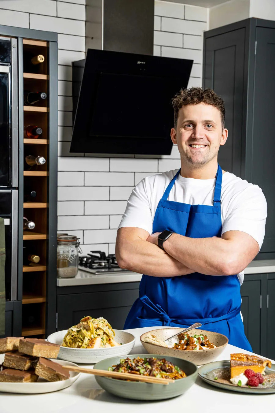 Jon Watts wearing a blue apron standing in a modern kitchen